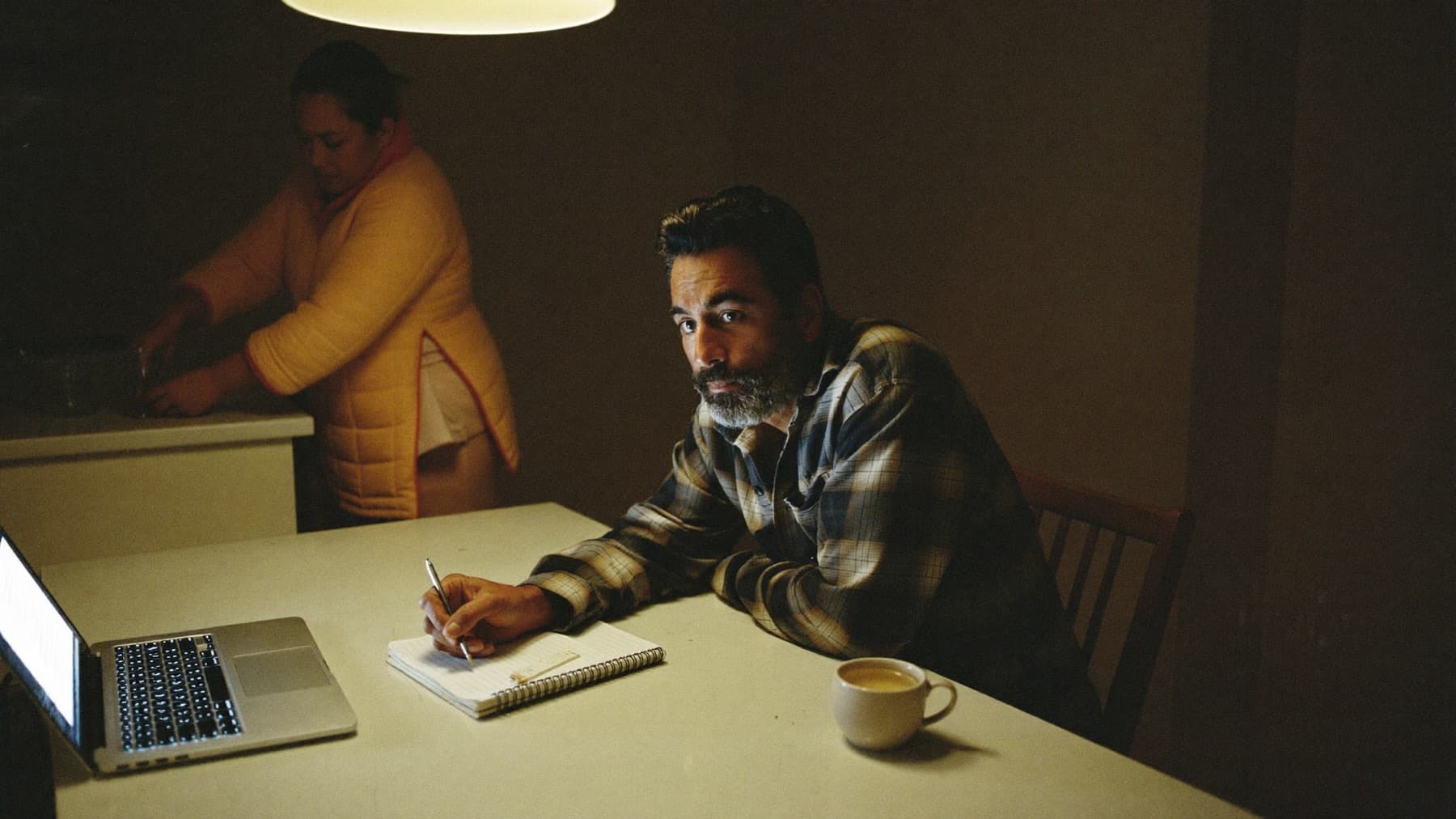 A man sits at a kitchen table late at night with a spiral notebook, a pen and an open laptop, while behind him in the dim kitchen someone in a quilted dressing gown stands at the counter.