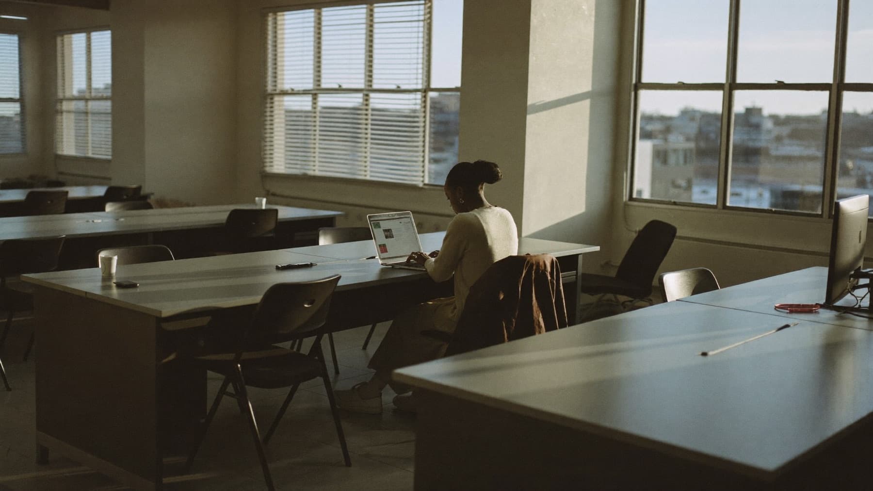A woman works alone at a laptop in a wide open-plan office at the end of the day, the desks and chairs around her cleared and empty, her coat over the back of her seat.