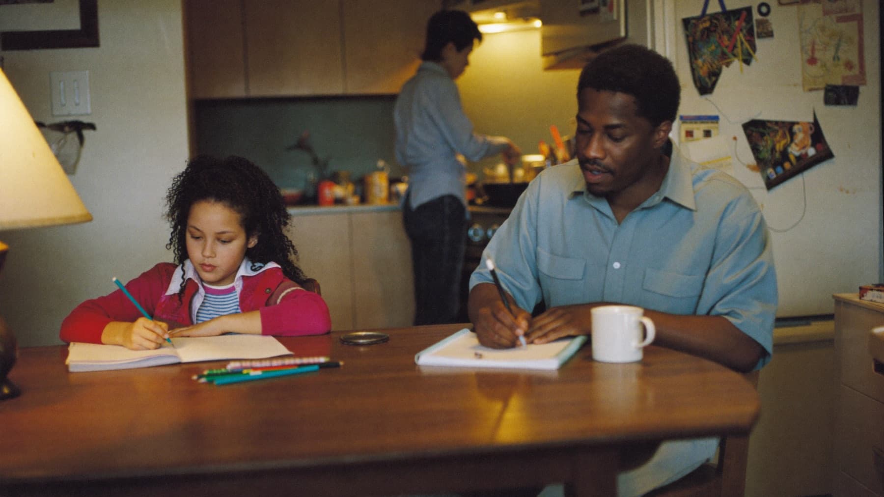 A father writes in a notebook at a kitchen table on an ordinary evening while his young daughter does homework beside him and a parent cooks at the stove behind them, children's drawings taped to the fridge.