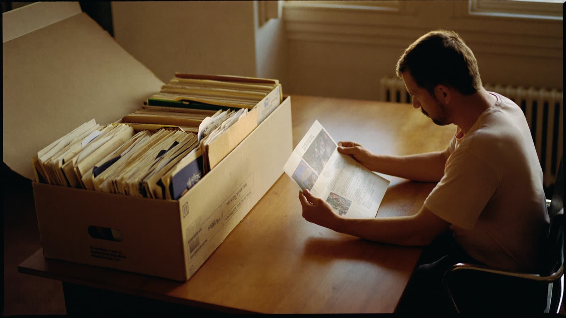 A man at a desk beside an open archive box crammed with old project folders, reading a single thin printed page.
