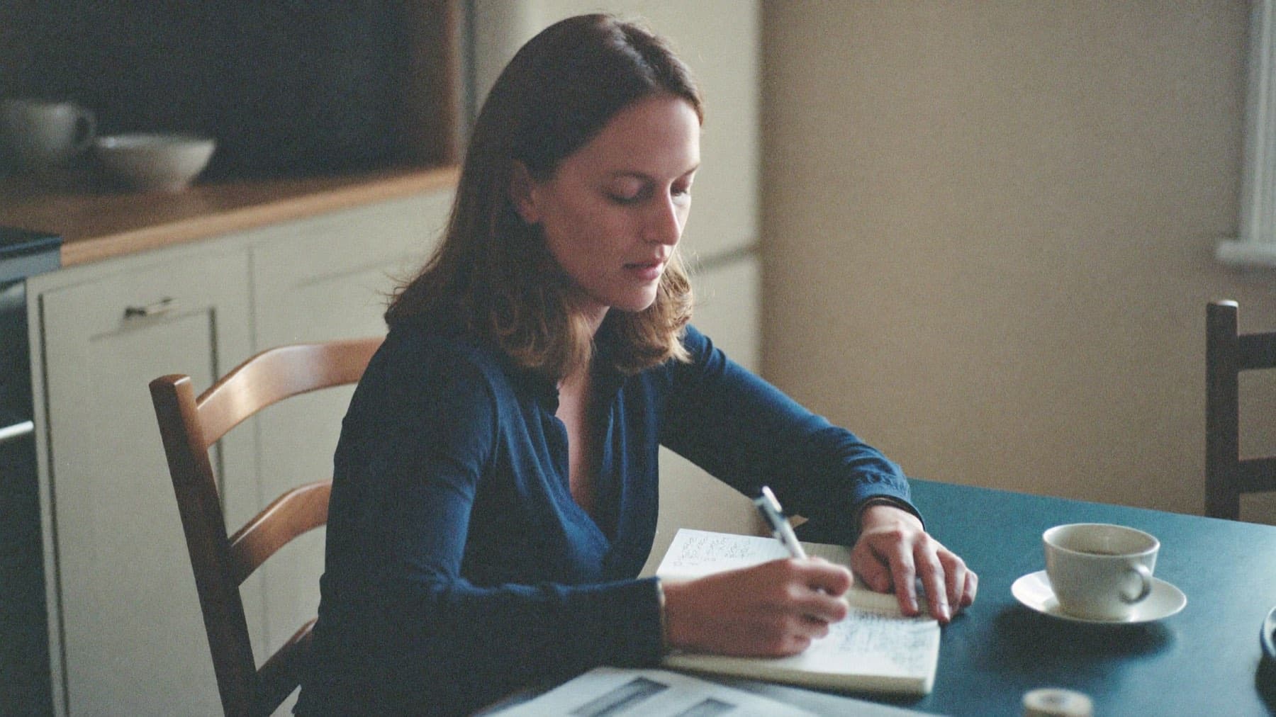 A woman at a kitchen table in early morning light, writing carefully in a notebook beside a closed laptop and a cup of coffee.