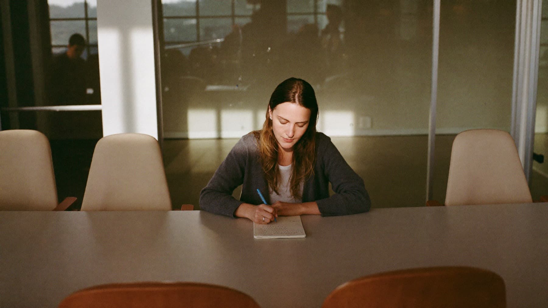 A woman alone in a glass-walled meeting room just after everyone has left, writing a few lines in a notebook while the chairs are still pushed out.