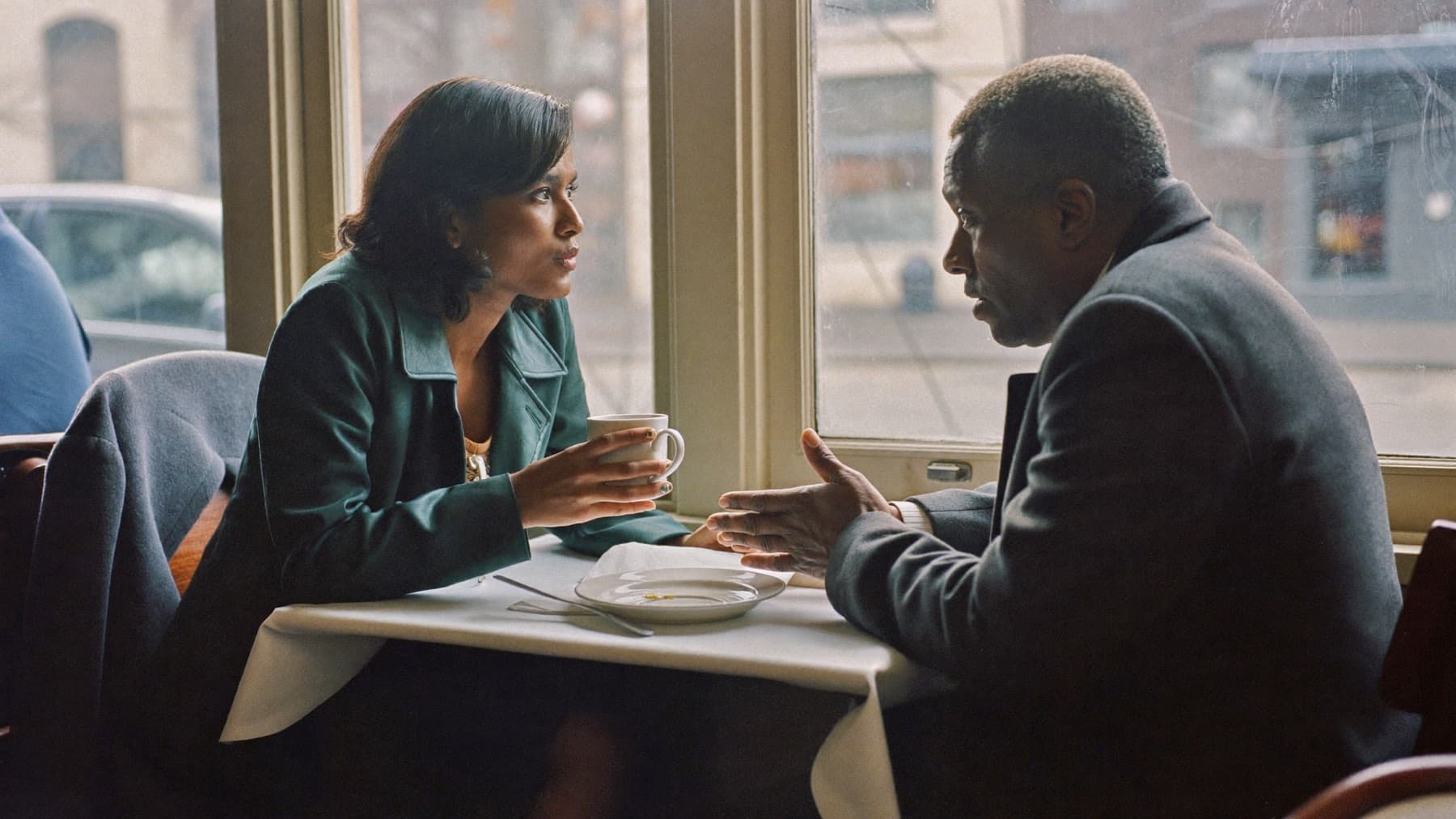 Two people in coats talk across a small café table by a window on a grey afternoon, one holding a mug with both hands and the other mid-gesture, an empty plate between them.