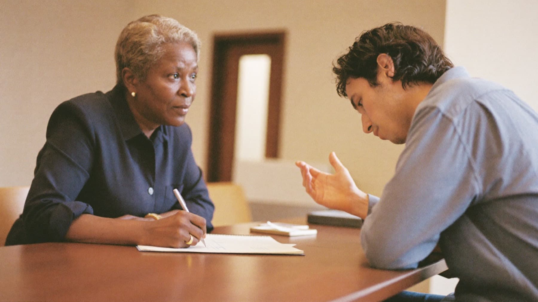 An interviewer sits with her pen paused above her notepad, listening closely to a candidate who is mid-answer.