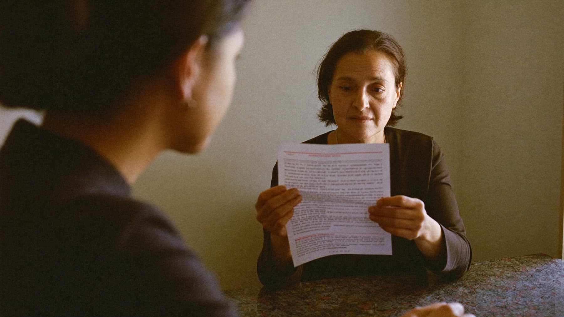 An interviewer reads a single printed page closely across a table while a candidate sits opposite.