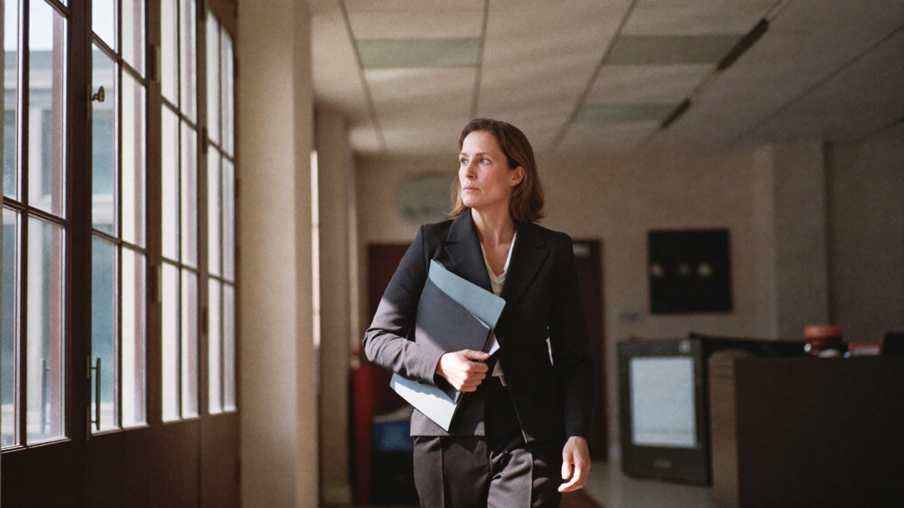 A composed professional walking through a sunlit corridor, carrying a folder.