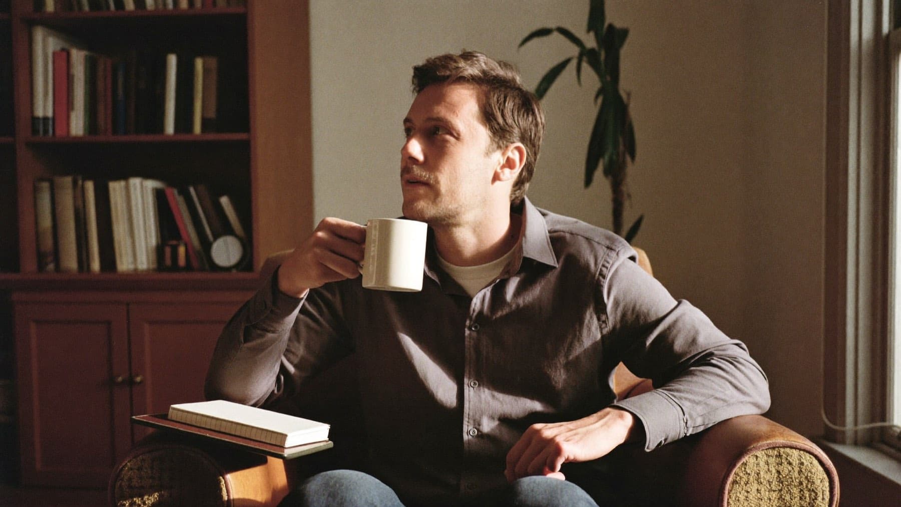 A man sitting in an armchair by a window with a mug of coffee, thinking something over.