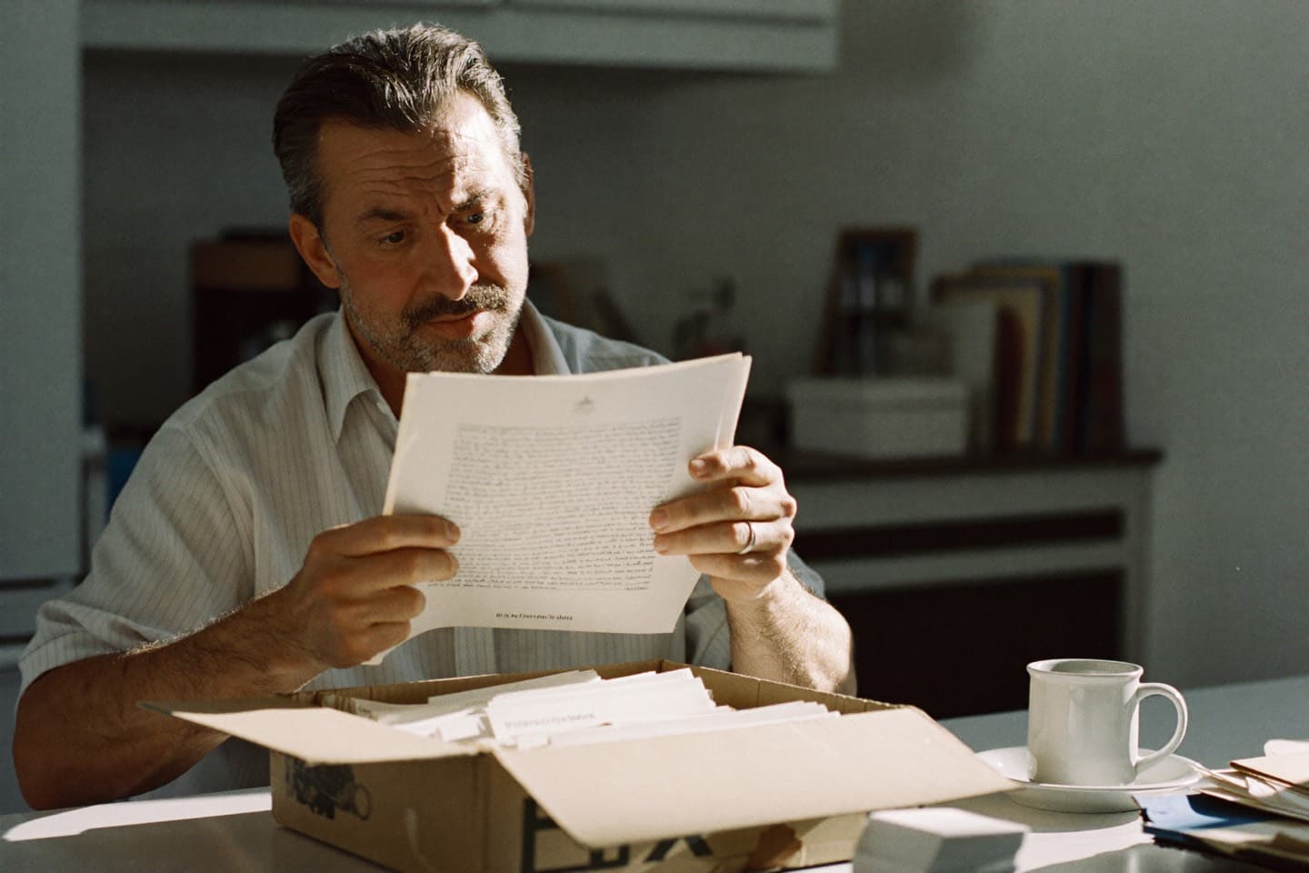 A man at a kitchen table in morning light, methodically reading through a document from a box of old work papers.