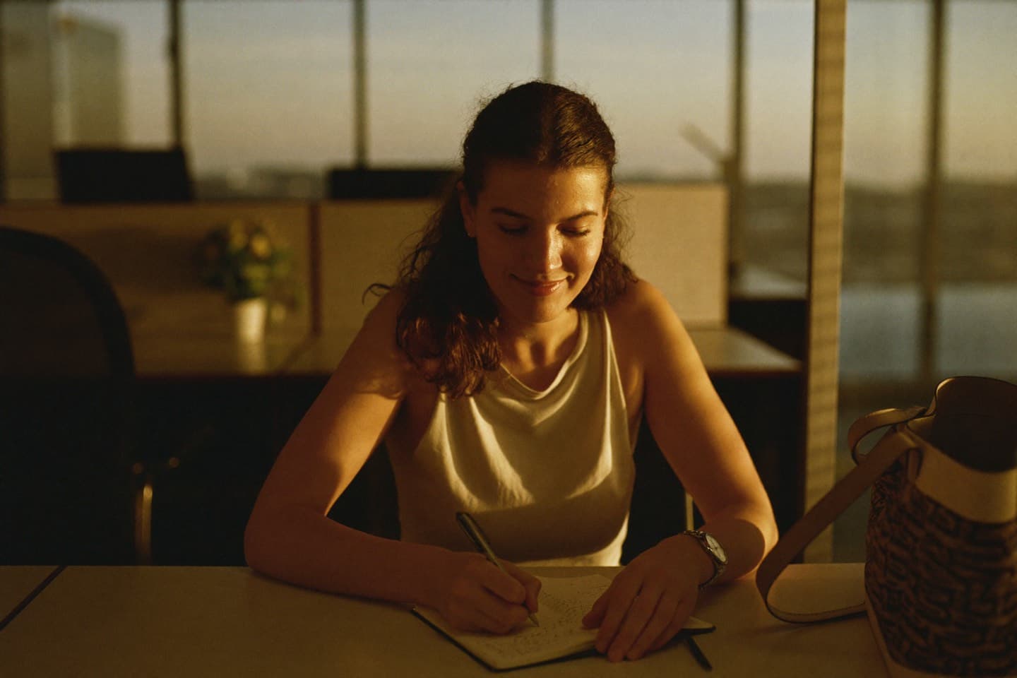 A woman at her desk in warm evening light, writing a few lines in a notebook before heading home.