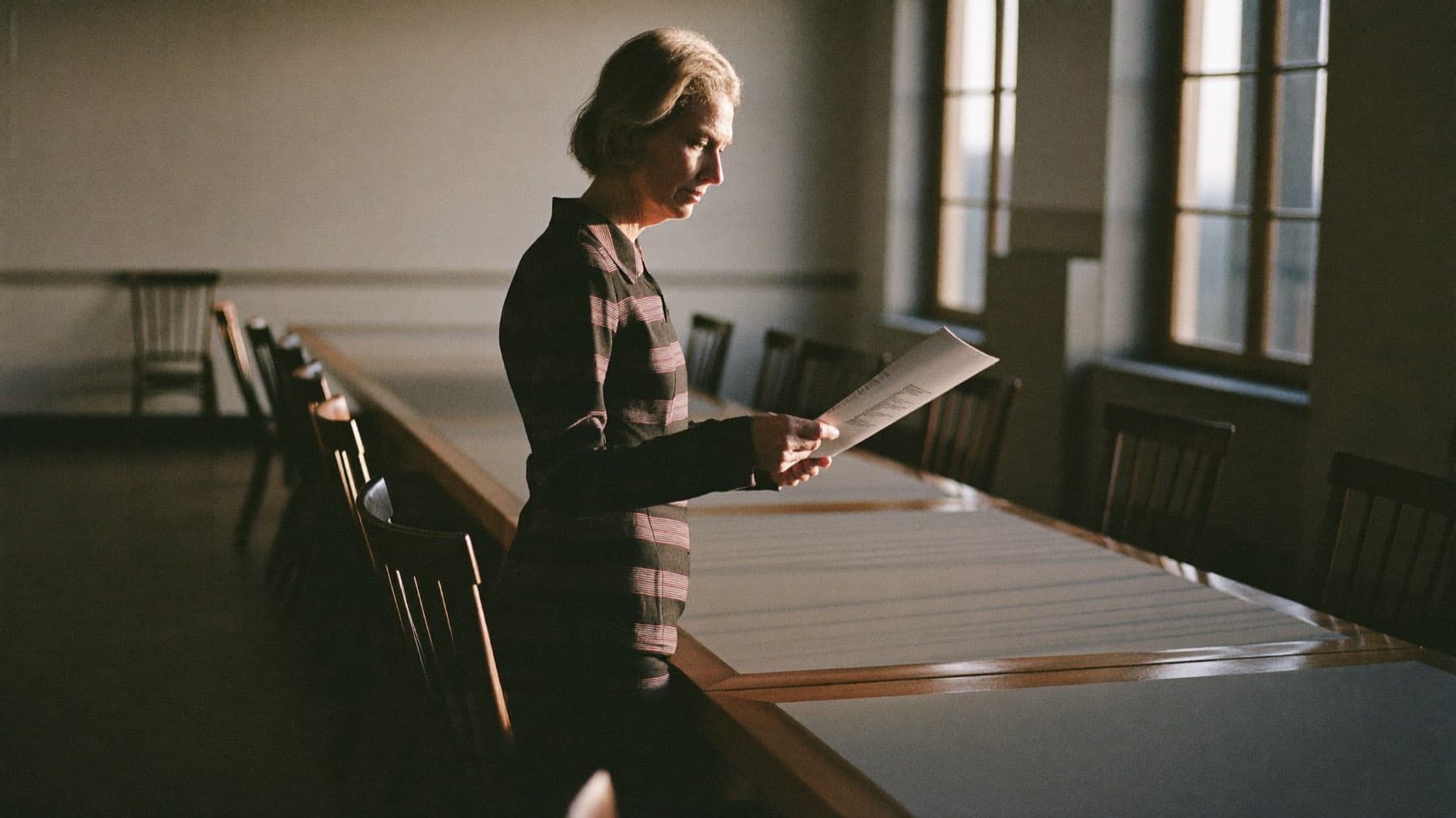 A senior executive standing beside a long boardroom table in late-afternoon light, reading a printed document.
