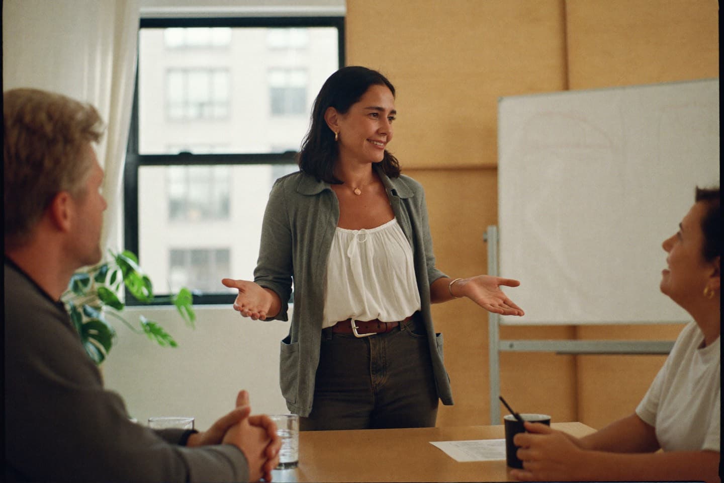 A woman standing at the head of a small meeting table, mid-sentence, leading a discussion with two colleagues.