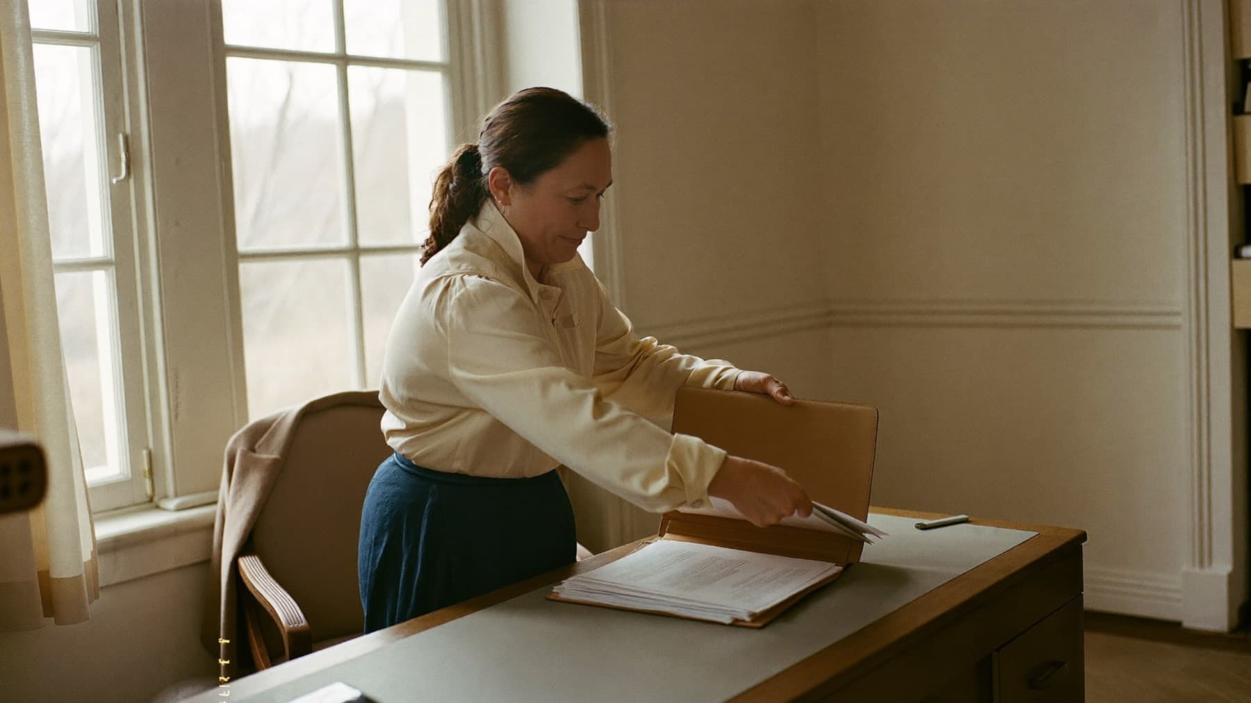 A woman standing at a cleared desk by a tall window in soft morning light, calmly placing her printed pages into a leather folder she is about to carry out.