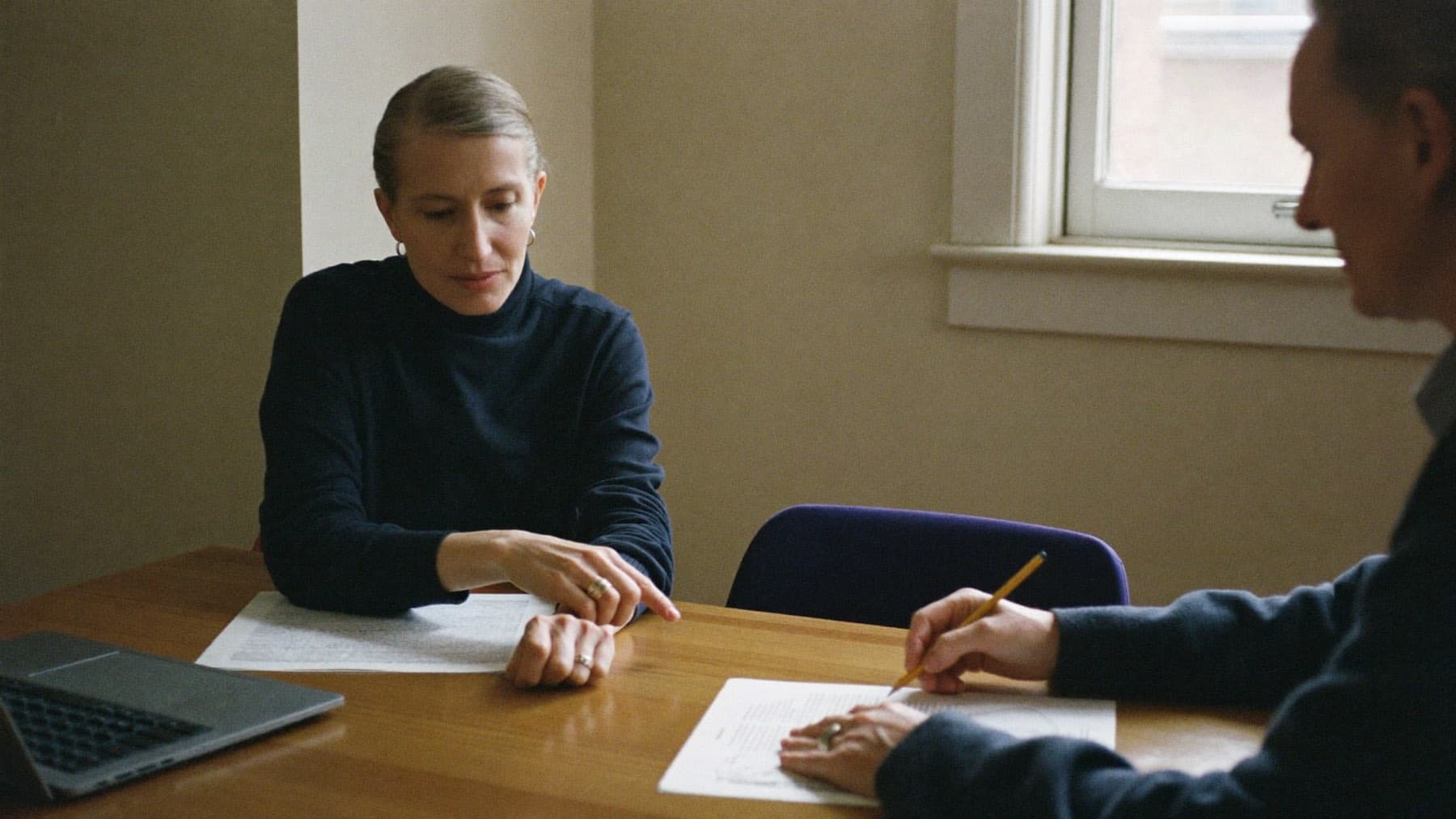 A career coach and her client sitting at a wooden table in a quiet office, both working over the same printed pages, one pointing at a line and the other writing a note in the margin.