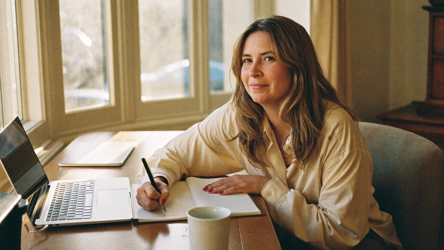 A woman writing in a notebook at her desk in warm afternoon light, unhurried.