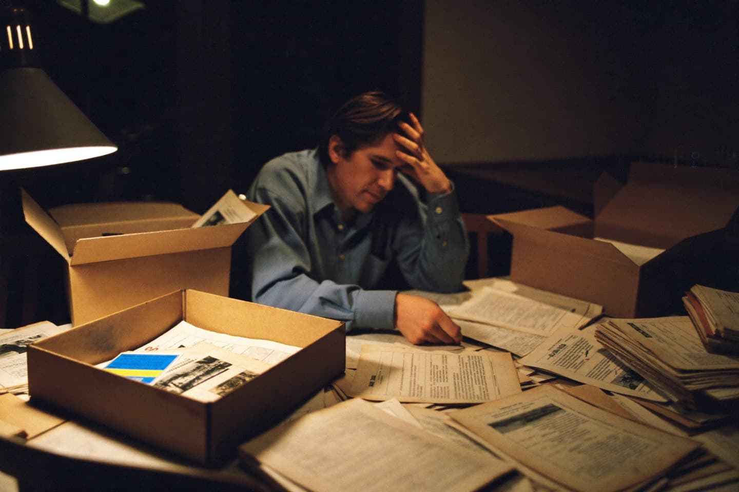 A man late at night surrounded by scattered documents and open file boxes, trying to reconstruct old work.