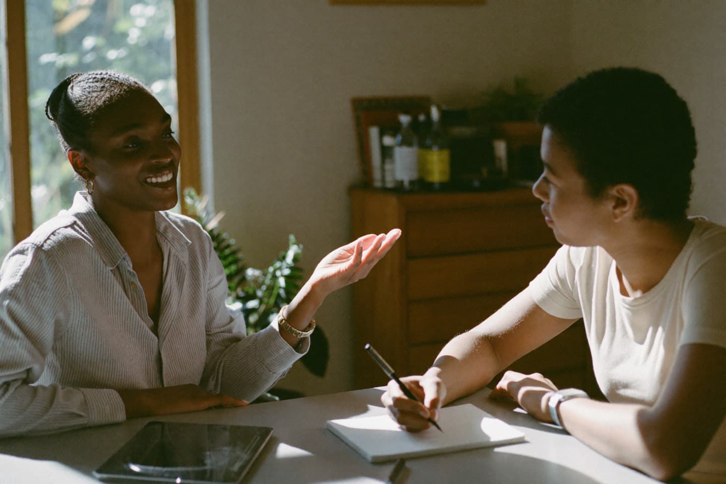 Two people talking across a table in a sunlit room, one speaking easily while the other takes notes.
