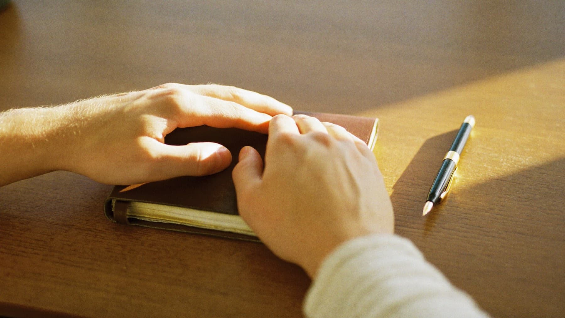 A pair of hands resting on a well-worn personal notebook on a desk in warm morning light, a pen beside it.
